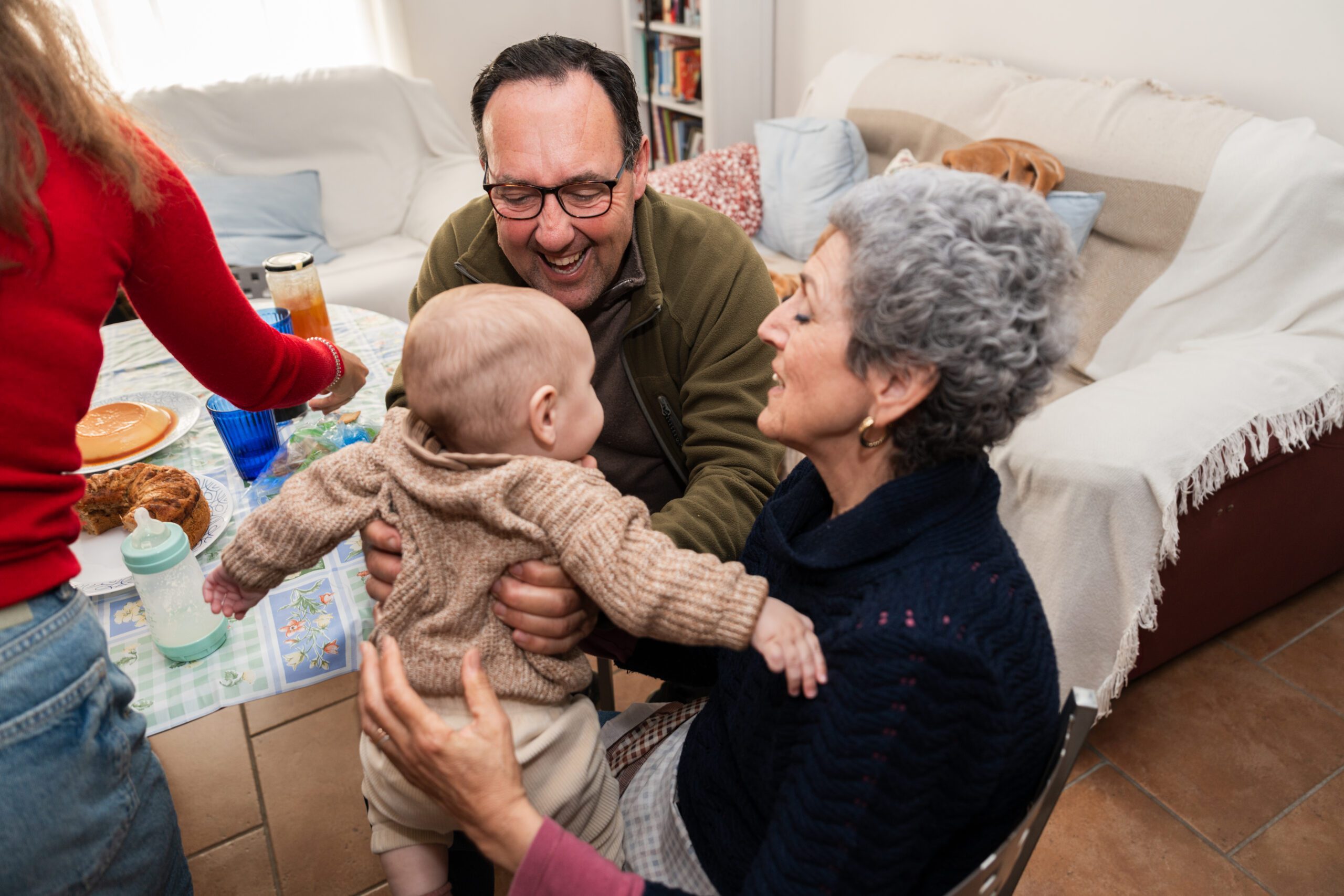 Grandparents holding their grandchild while the family is gathering around the table for a meal